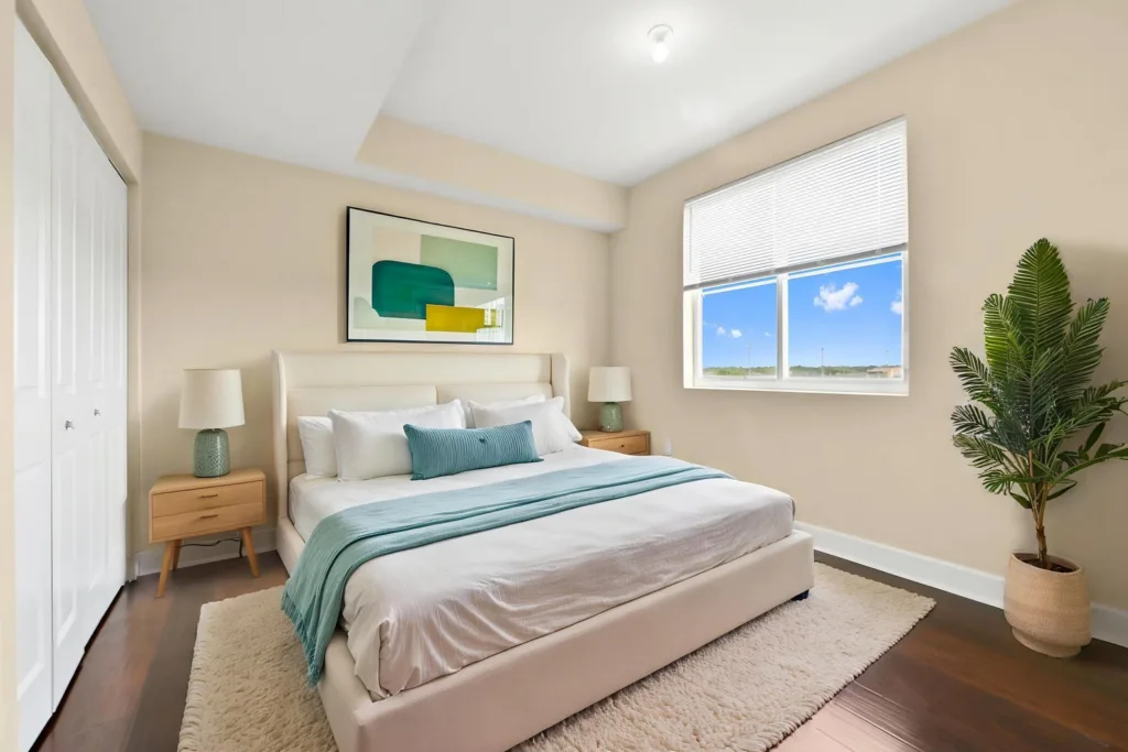 20261007214752721548000000-o - Lake Worth Homes - Lake Worth Homes Bright, modern bedroom with a cream upholstered king bed, teal throw and pillows, and wooden nightstands on either side under beige walls and a window view.