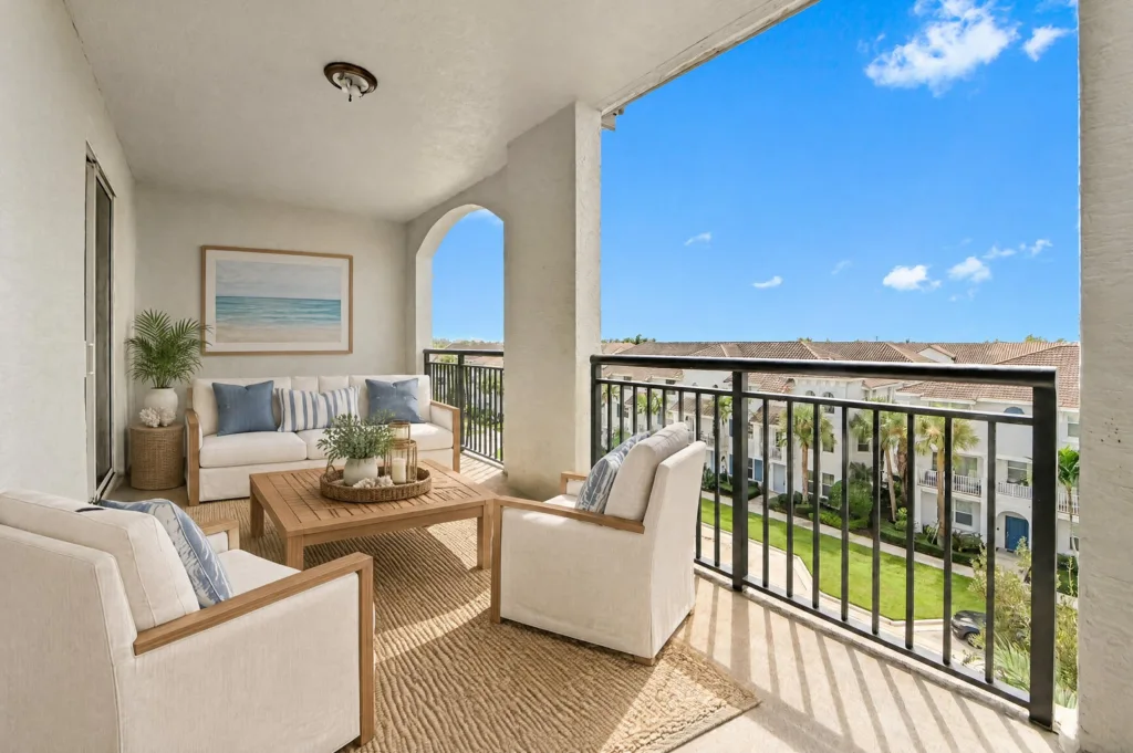20261007214958087589000000-o - Lake Worth Homes - Lake Worth Homes Cozy balcony seating area with cream cushions and blue pillows, wooden coffee table, and potted plant, overlooking a sunny courtyard and blue sky.