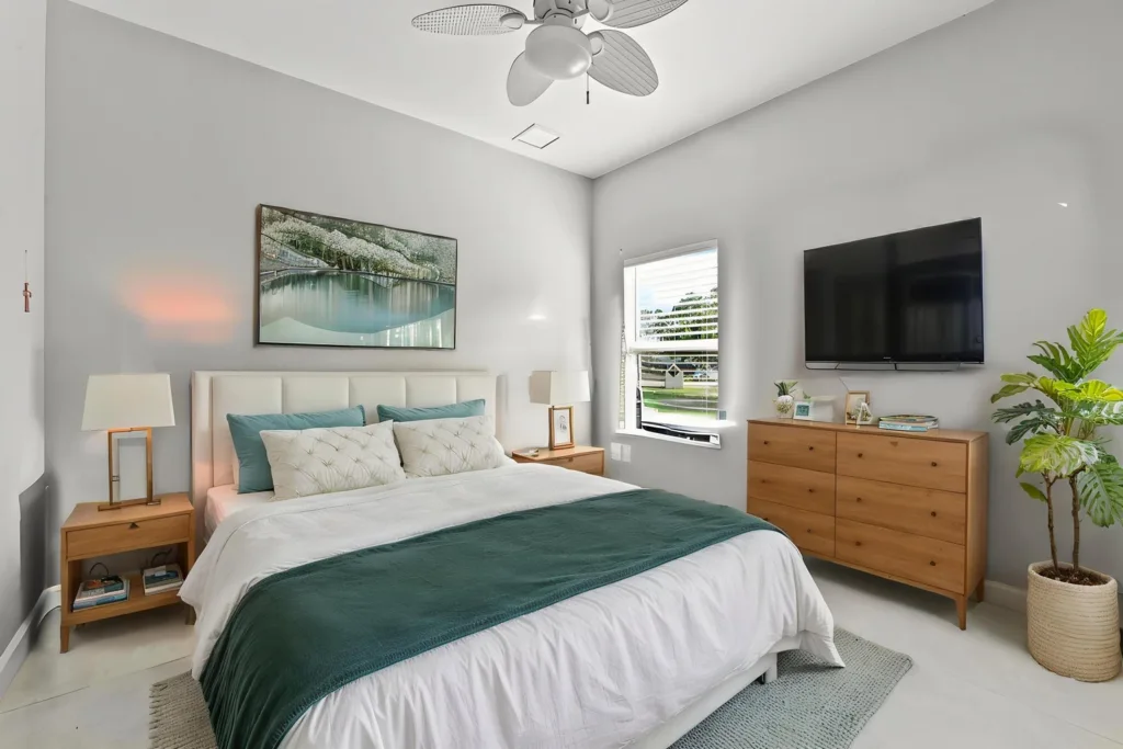 Modern bedroom with a white upholstered bed, teal pillows, two wooden nightstands with lamps, and a wooden dresser with a mounted TV above.