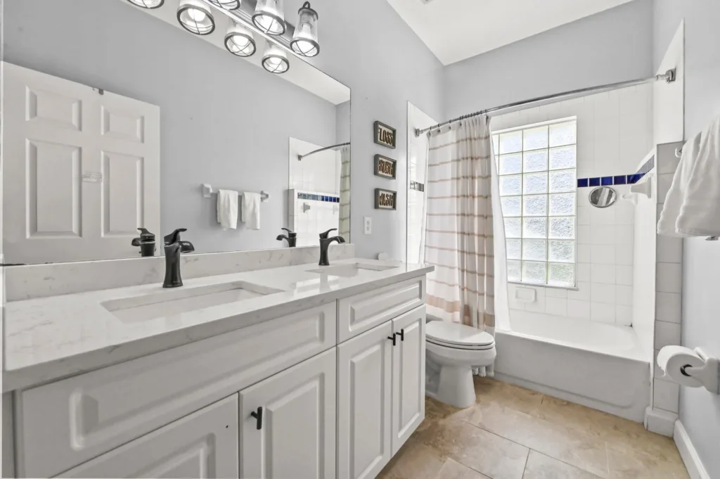 Bright white bathroom with a double vanity, marble countertop, and black faucets, large mirror and light fixture, plus a tub/shower with a striped curtain.