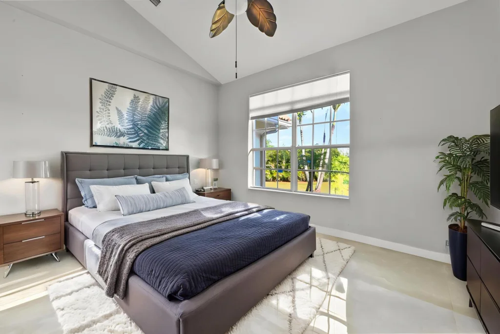 Bright modern bedroom with a gray upholstered king bed, two wooden nightstands, and a large window overlooking greenery outside.
