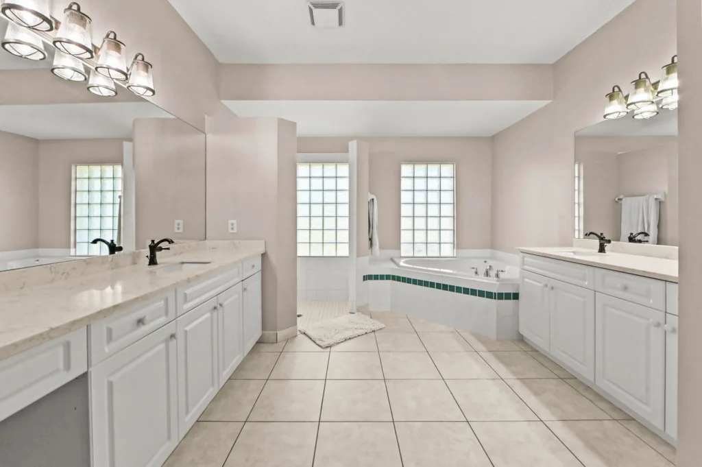 Bright bathroom with dual vanities, marble countertops, white cabinets, a corner tub with green tile trim, and glass block windows.