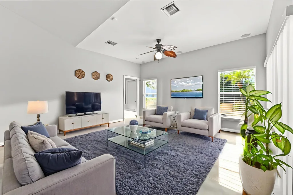Bright living room with gray walls, a three-seat sofa and two armchairs around a glass coffee table, TV on a white console, and a blue rug.