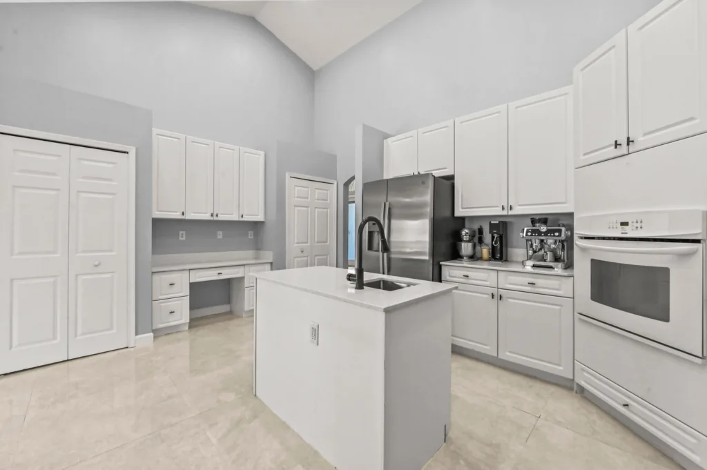 Modern white kitchen with a central island sink and black faucet, stainless fridge, and abundant cabinetry.
