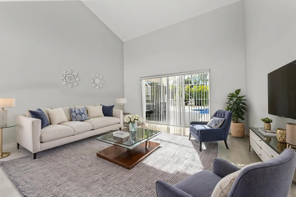 Bright living room with pale gray walls, white sofa with blue and cream cushions, glass coffee table, and a blue accent chair by a sliding-glass door with blinds.