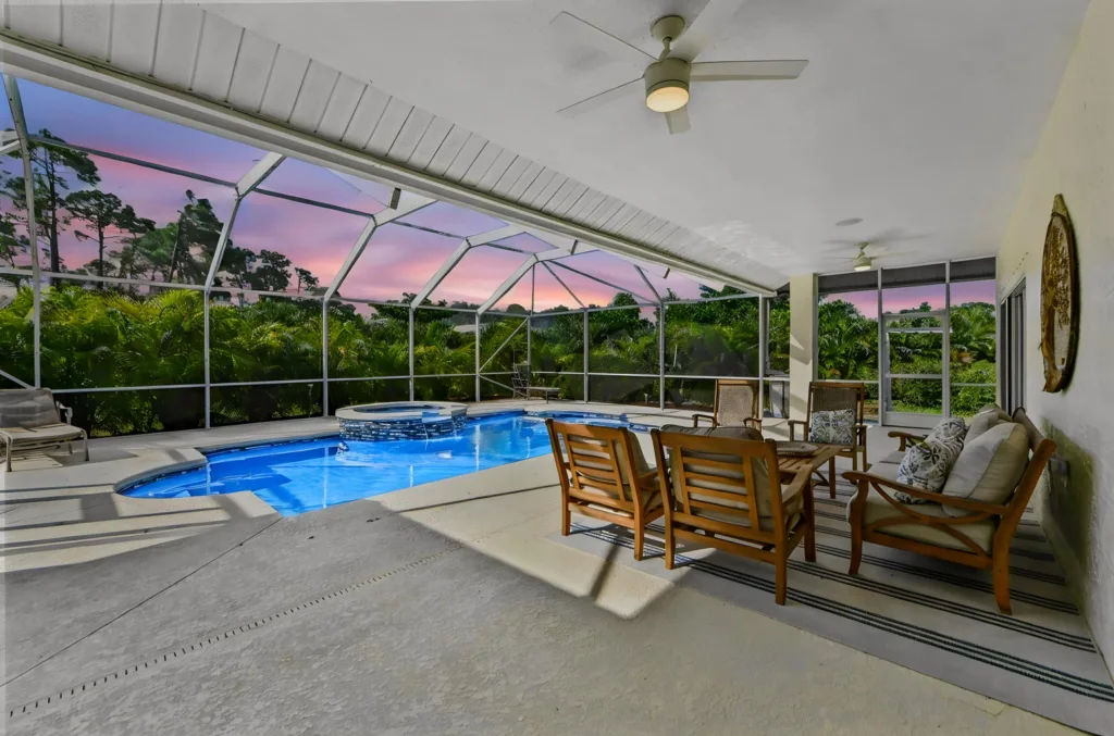 Screened pool deck with a blue curved pool, wicker seating, and tropical trees at sunset outside the enclosure.
