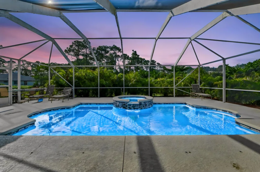 Screened-in backyard pool with a raised circular spa in the center, surrounded by lounge chairs and tropical greenery at sunset.