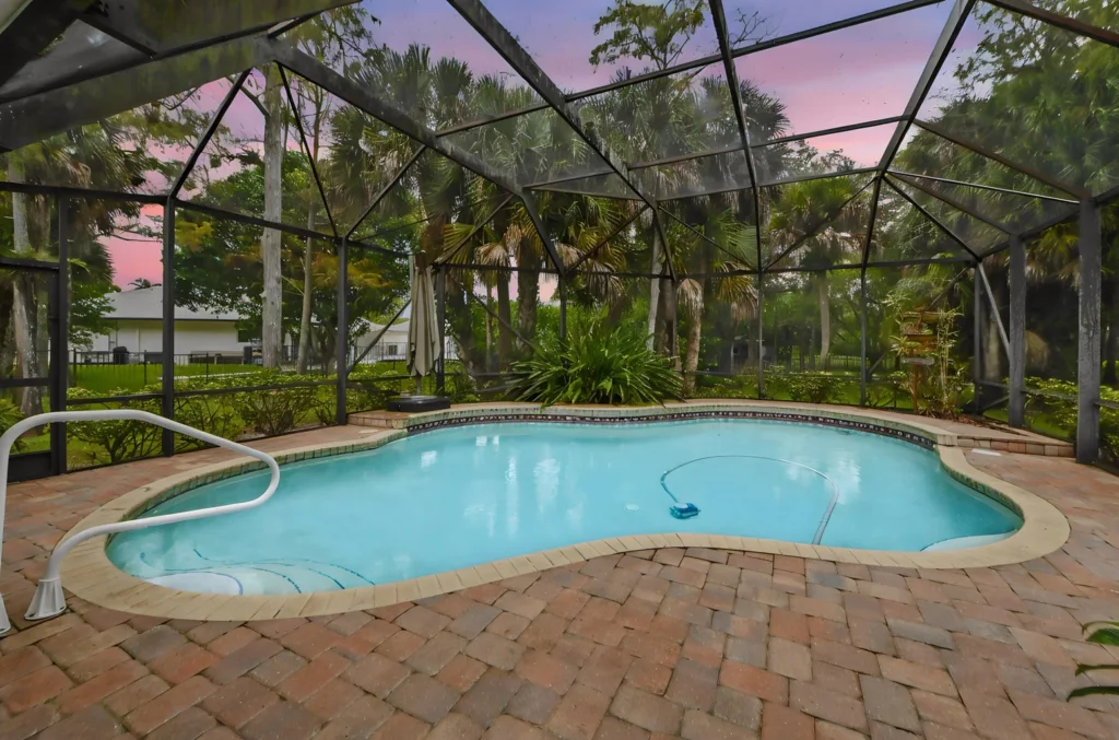 Screened-in backyard pool with curved edges, brick paver deck, and tropical plants, sunset pink sky in the background