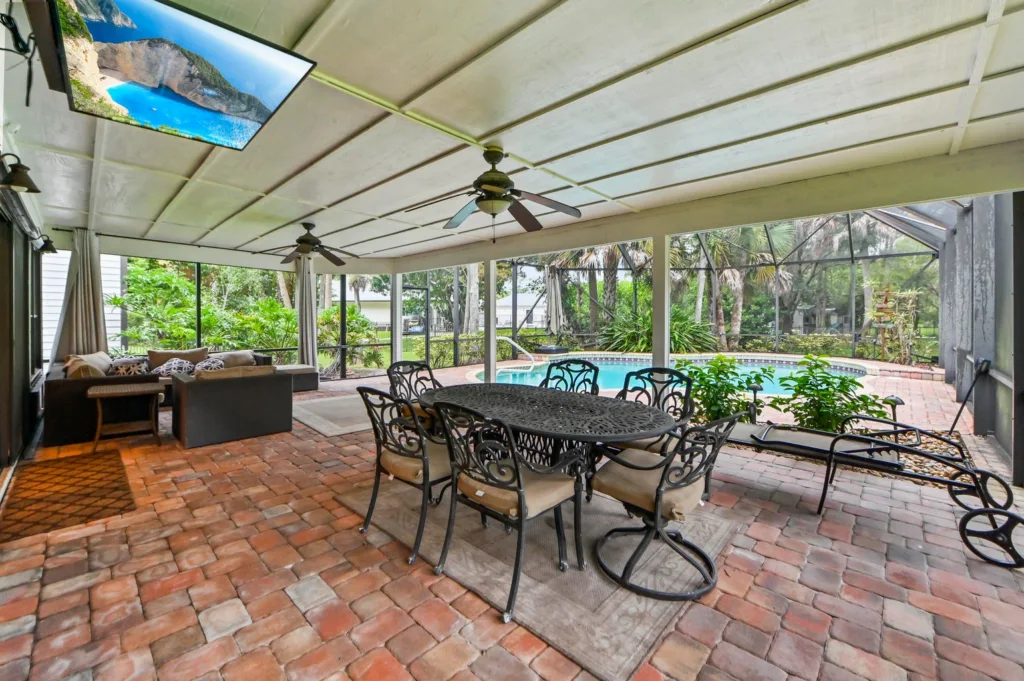 Covered outdoor living area with brick paver floor, a metal dining table and eight cushioned chairs overlooking a screened backyard pool.