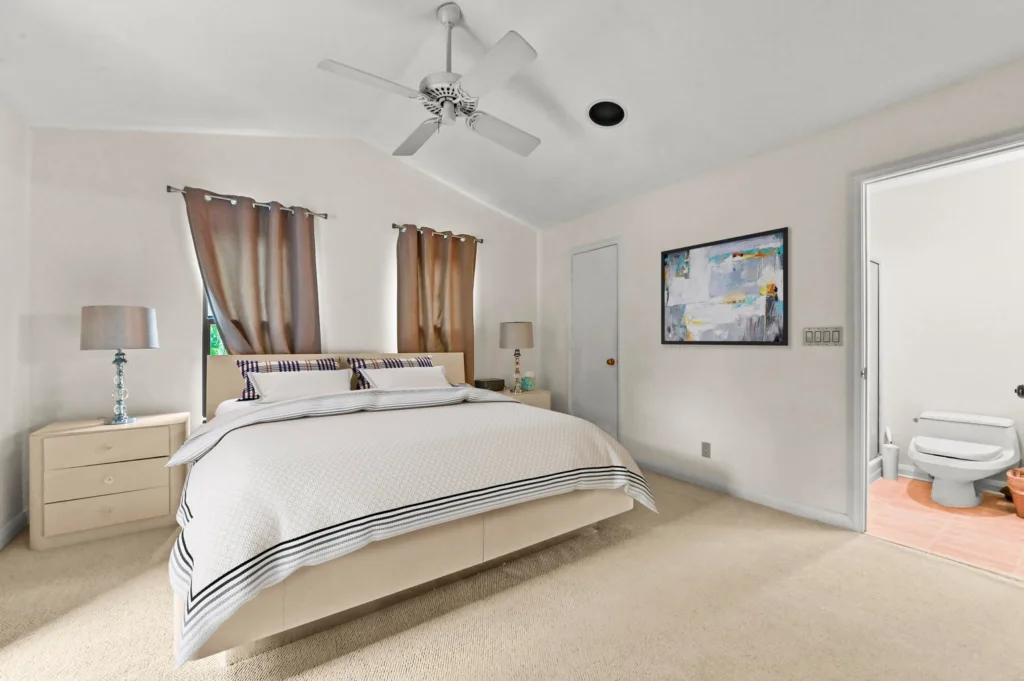 Bright beige bedroom with a king bed, two nightstands, and matching lamps; brown curtains and abstract wall art, plus an adjacent pink-tiled bathroom with a toilet.