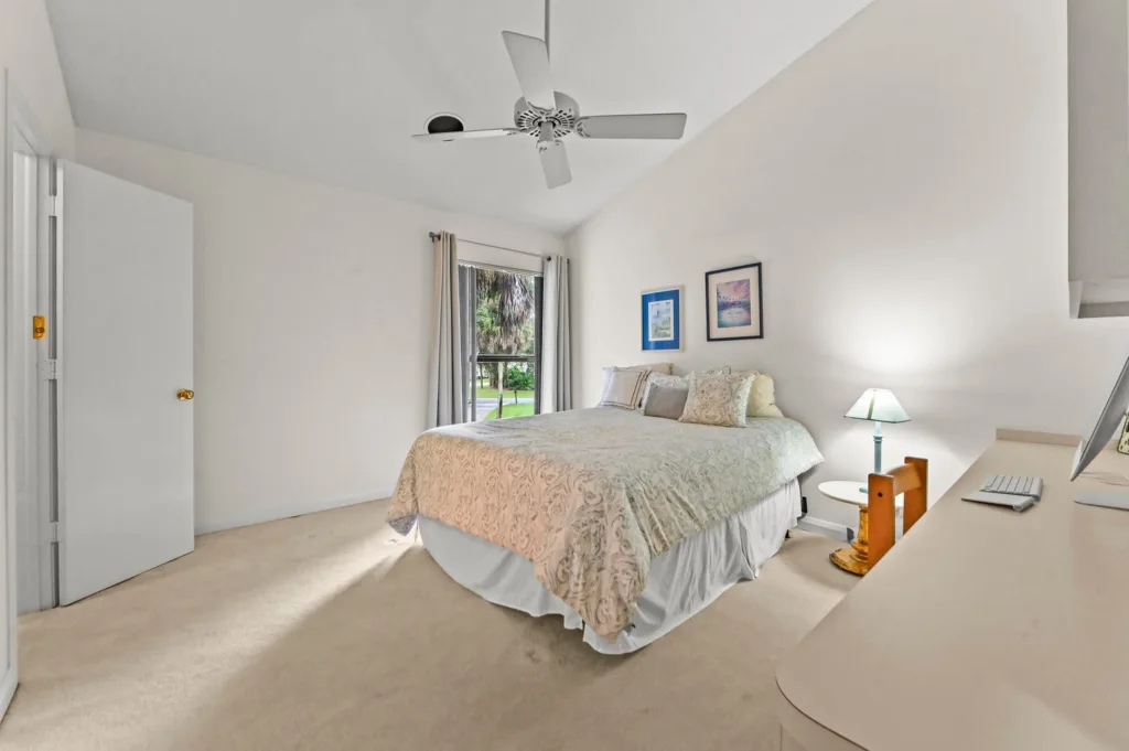Bright bedroom featuring a large bed with a beige patterned cover, multiple pillows, and a white bed skirt, near a window with light curtains and a ceiling fan overhead. Desk area on the right shows a lamp, keyboard, and monitor, suggesting a small home office setup in a calm, neutral room.