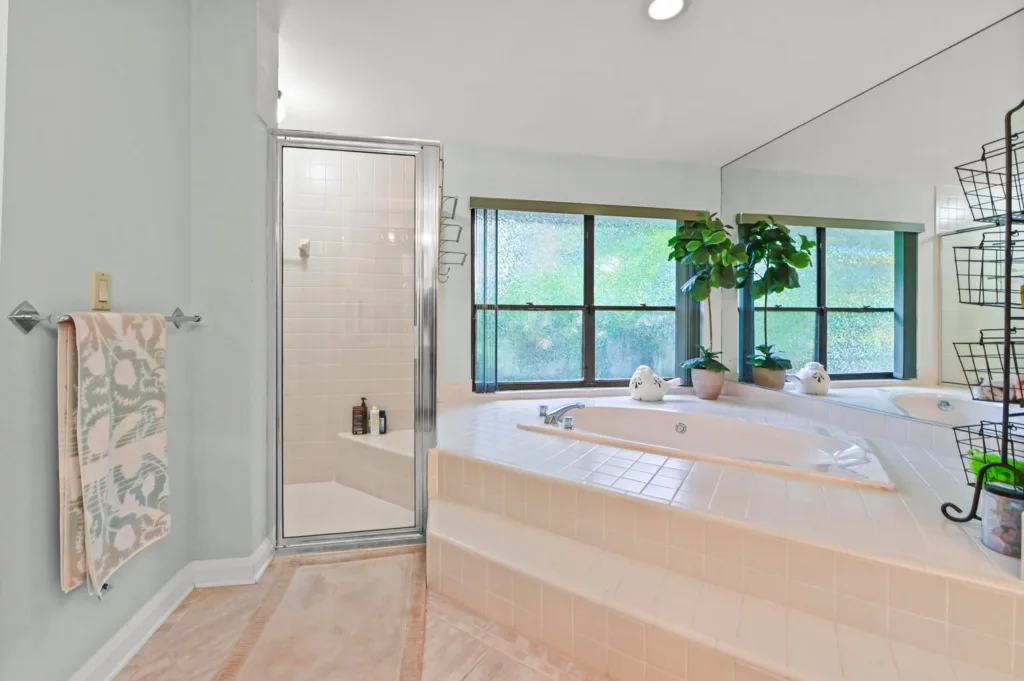 Bright bathroom with a corner glass-enclosed shower and a large tiled soaking tub by a window, topped with green potted plants and towels nearby.