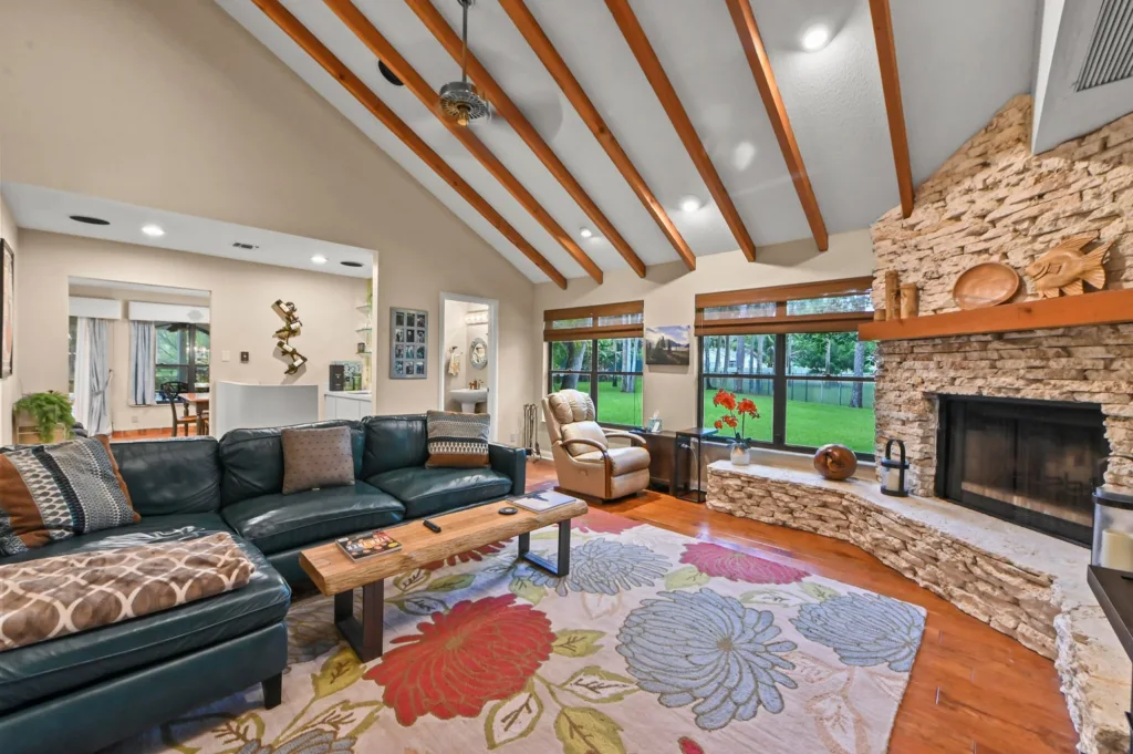 Cozy living room with a dark leather sectional, colorful floral rug, and a stone fireplace beside large windows showing a green backyard.