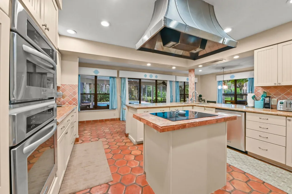 Bright kitchen with a central island, stainless steel range, and a large industrial vent hood; terracotta tile floor and blue-curtained windows.