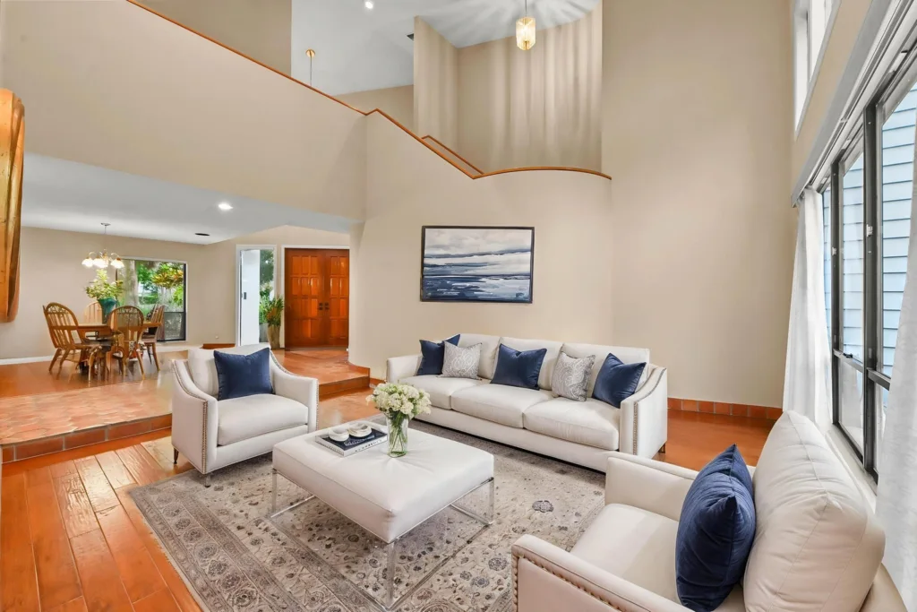 Spacious living room with white sofas and navy throw pillows, a glass-tinished coffee table, and a patterned rug, with large windows to the right and a dining area in the back left.
