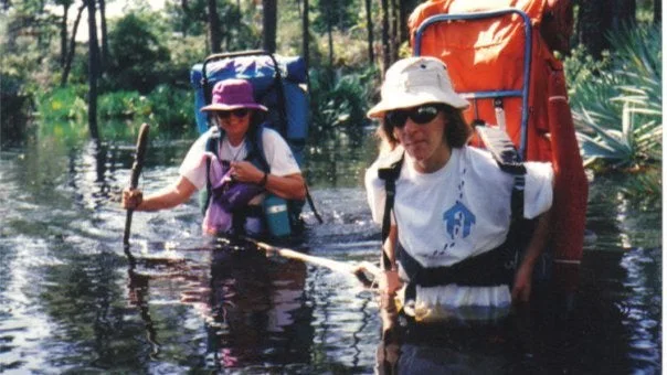 Two hikers wading through knee-deep water in a flooded forest, each wearing large backpacks and hats.