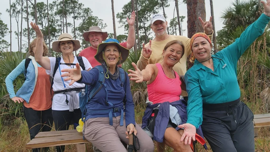 Group of smiling hikers posing with arms raised in a sunny outdoor trail setting with trees and grasses behind them.