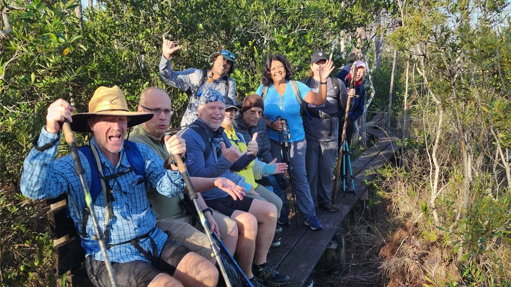 Group of hikers on a wooden boardwalk in a forest, smiling and waving at the camera.