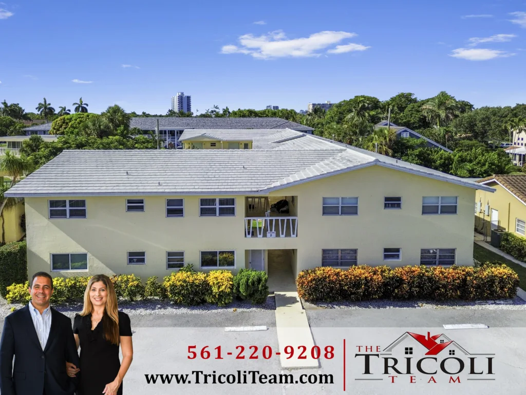 Two smiling real estate agents in business attire stand in front of a pale yellow apartment building with hedges, and a large Tricoli Team banner with a phone number at the bottom.