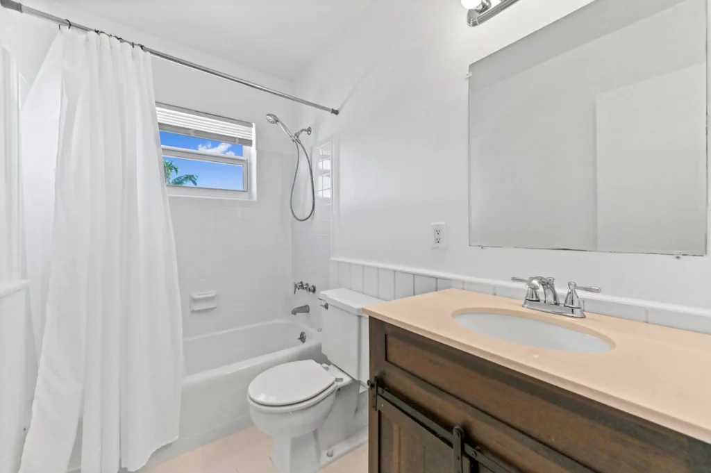 White bathroom with a tub–shower combo, shower curtain, and a window showing blue sky; beige countertop vanity and mirror on the wall.