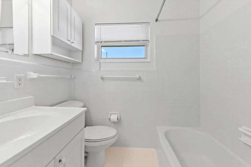 Small white bathroom with sink vanity, toilet, and bathtub; window with blinds and tiled walls.
