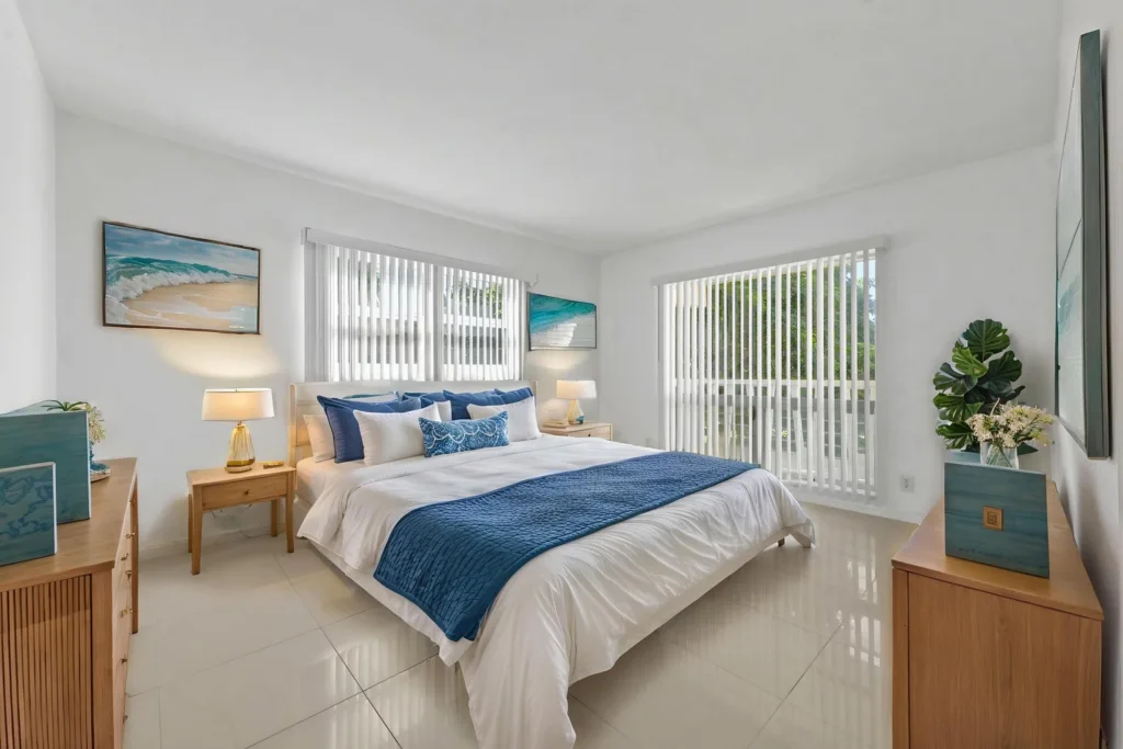 Bright coastal bedroom with white walls, a white bed, blue throw, and coastal artwork; large windows with vertical blinds and wooden nightstands.