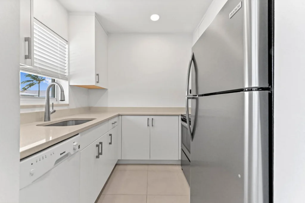 Compact white kitchen with beige countertops, a curved metal faucet over a sink by a window, and a stainless steel refrigerator on the right.