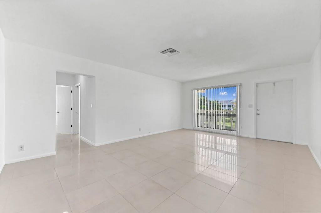 Bright empty living room with white walls and glossy tile floor, sliding glass door with vertical blinds opening to a green outdoor area on the right.