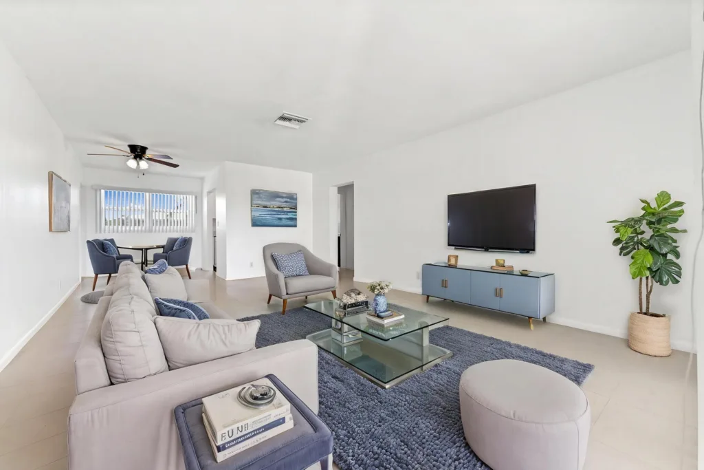 Bright, modern living room with a gray sectional, glass coffee table, blue rug, and a wall-mounted flat-screen TV. A tall plant sits by a blue console.