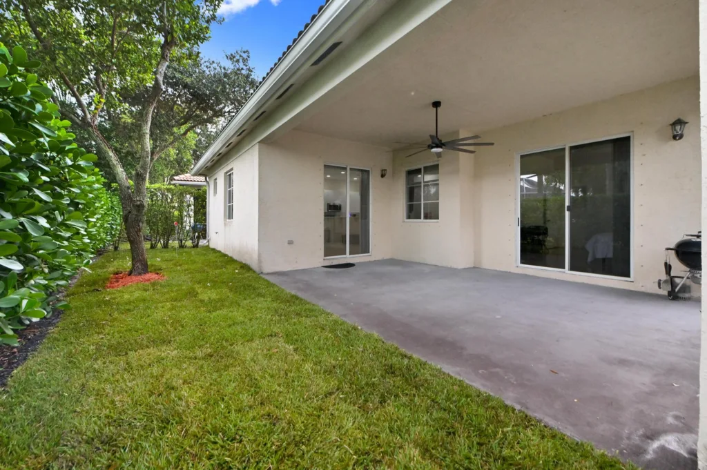 Backyard patio of a beige house with sliding glass doors, a ceiling fan, and a green lawn bordered by a hedge.