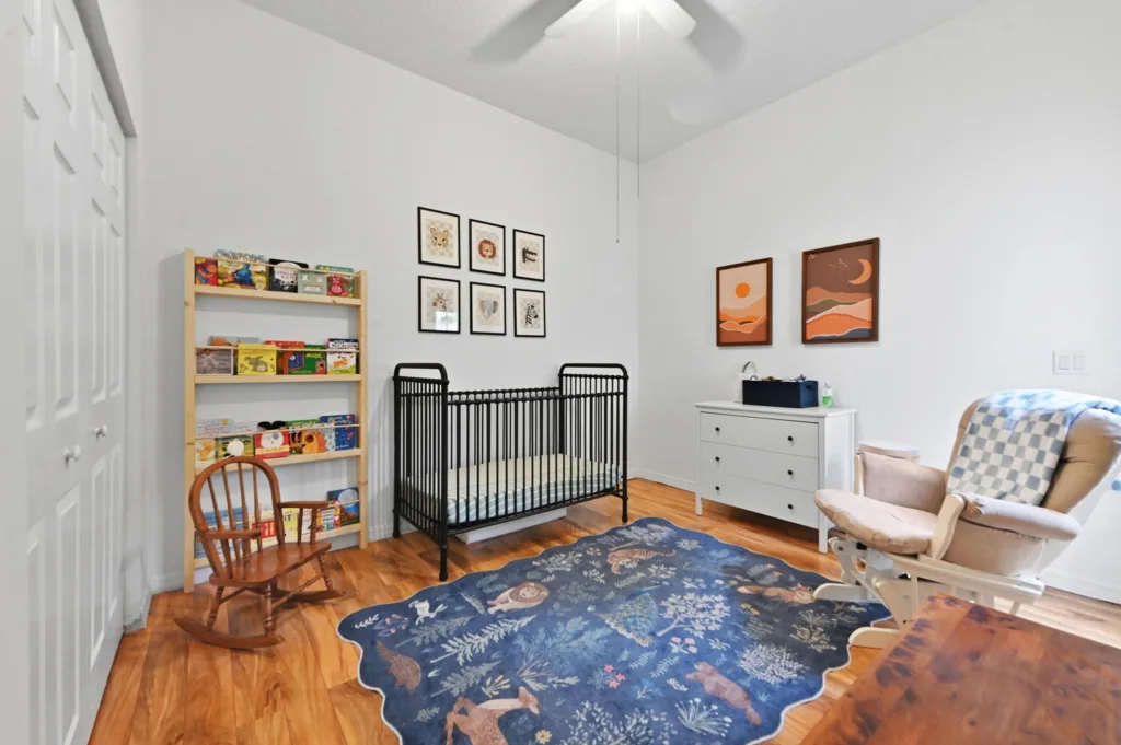 Nursery with a black metal crib, light wood bookshelf full of toys, white dresser, rocking chair, and blue animal-themed rug