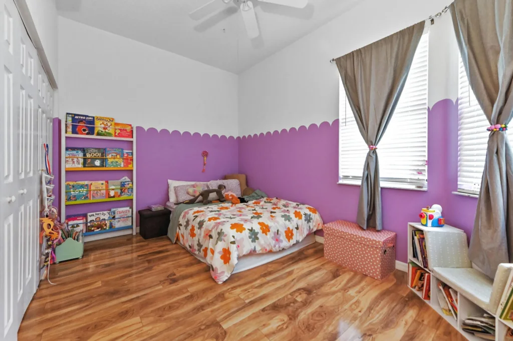 Child's bedroom with purple wainscoting, bed with floral quilt, and a tall white bookshelf filled with colorful books and toys on wooden floors.