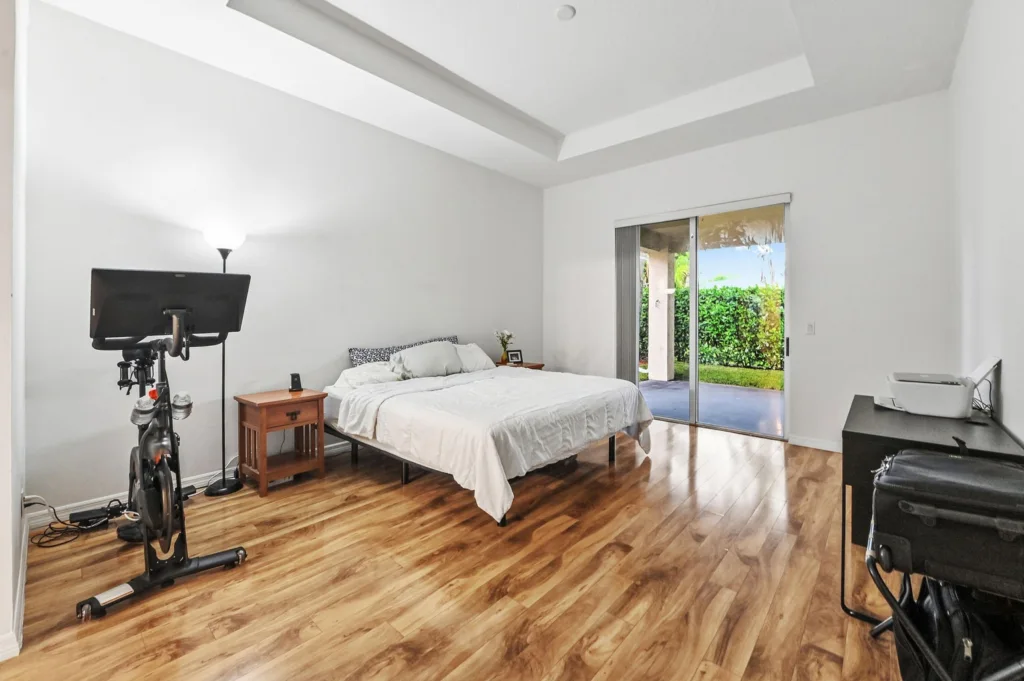 Bright bedroom with a stationary bike, queen bed, wooden nightstand, and sliding glass door to a garden.