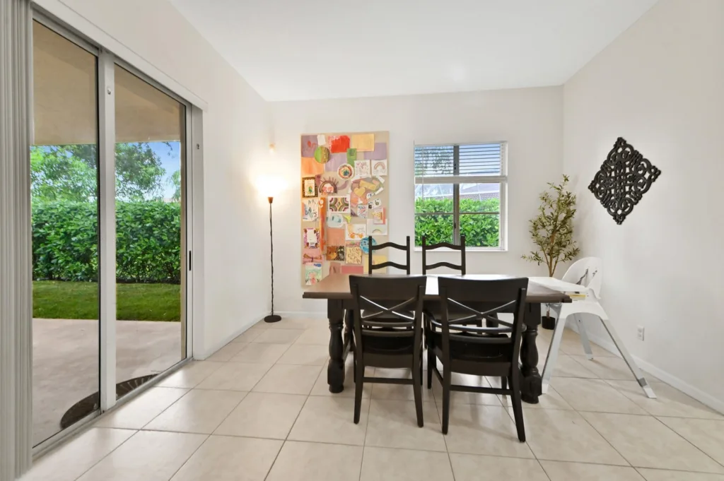 Bright dining room with a dark wood table and six chairs; sliding glass door to a green backyard on the left, colorful collage wall art in the center, and a window with blinds on the far wall.