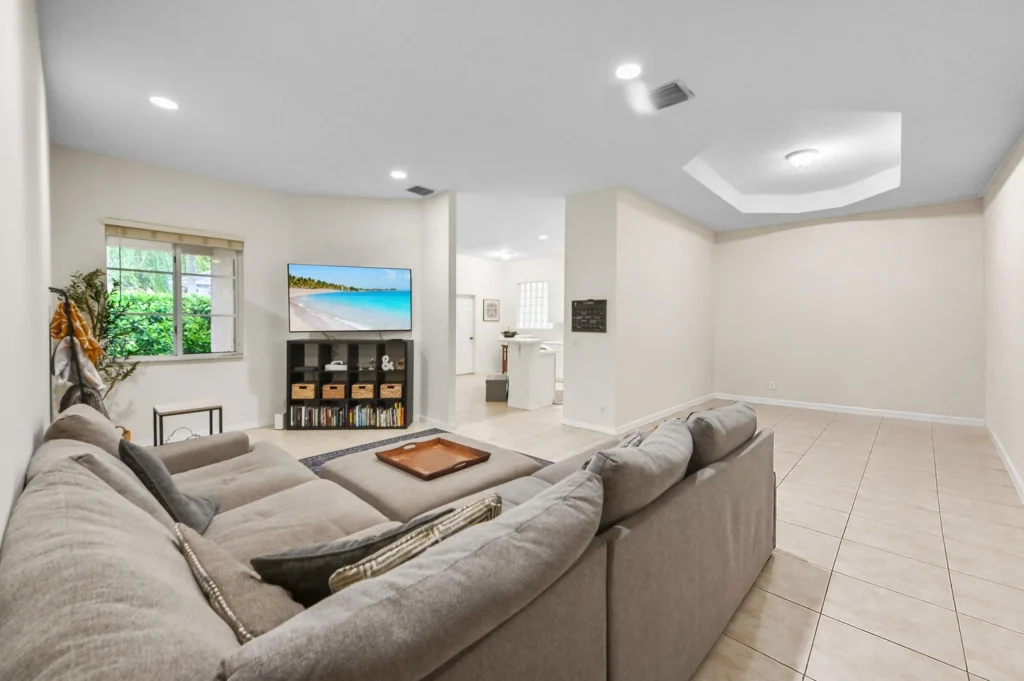 Open living room with a gray sectional sofa facing a flat-screen TV on a dark media console, window with greenery on the left, and tiled floor.