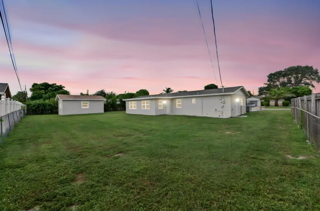 20261021192805520760000000-o - Lake Worth Homes - Lake Worth Homes Wide backyard at dusk showing a single-story gray house with a shed, green lawn, and fences along both sides under a pink-purple sunset sky