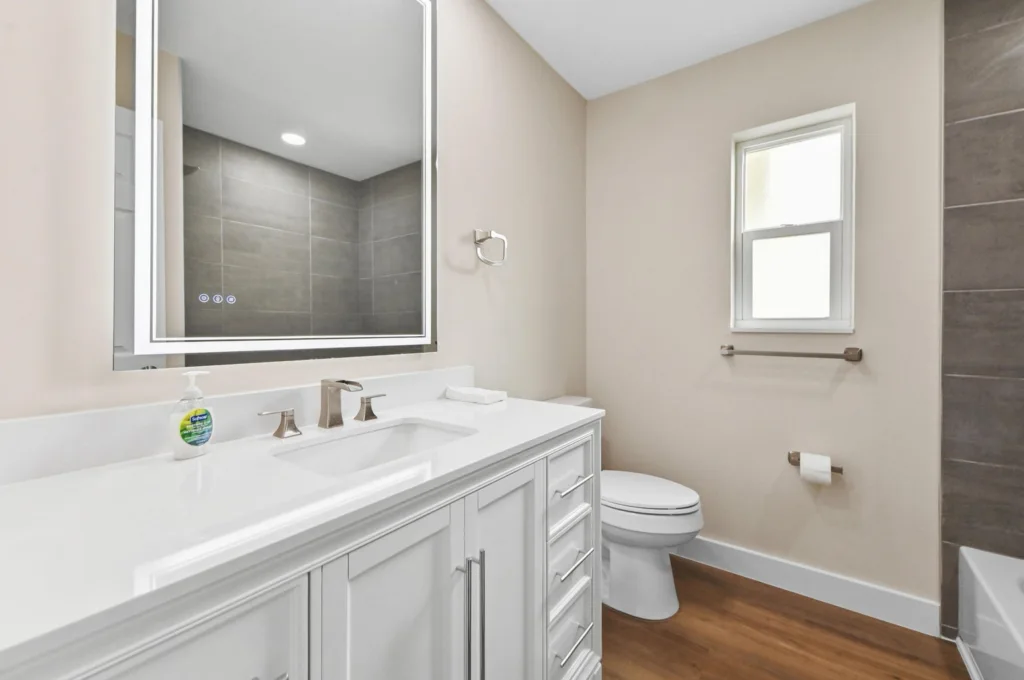 20261021192411144953000000-o - Lake Worth Homes - Lake Worth Homes Modern bathroom with white vanity and double sink, large backlit mirror, beige walls, wood-look floor, and a toilet by a window.