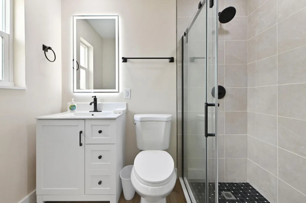 20261021192507002124000000-o - Lake Worth Homes - Lake Worth Homes Modern bathroom with white vanity, black faucet, backlit rectangular mirror, toilet, and glass-enclosed shower with black hardware.