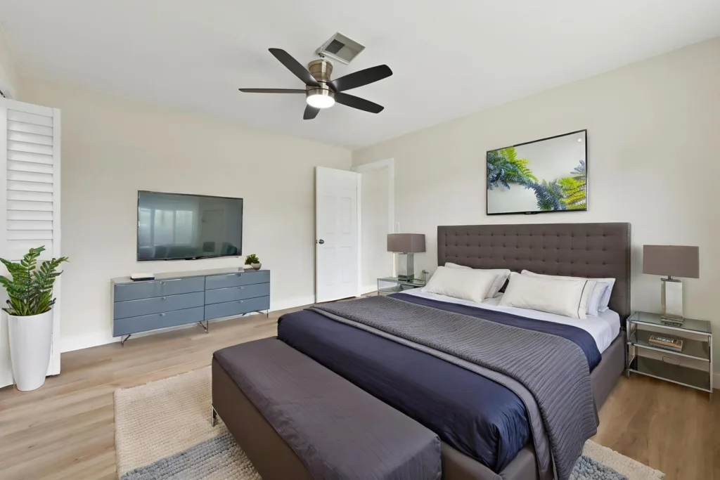 20261021192449541038000000-o - Lake Worth Homes - Lake Worth Homes Modern bedroom with a gray tufted king bed, nightstands, lamps, and a wall-mounted TV across from the bed.