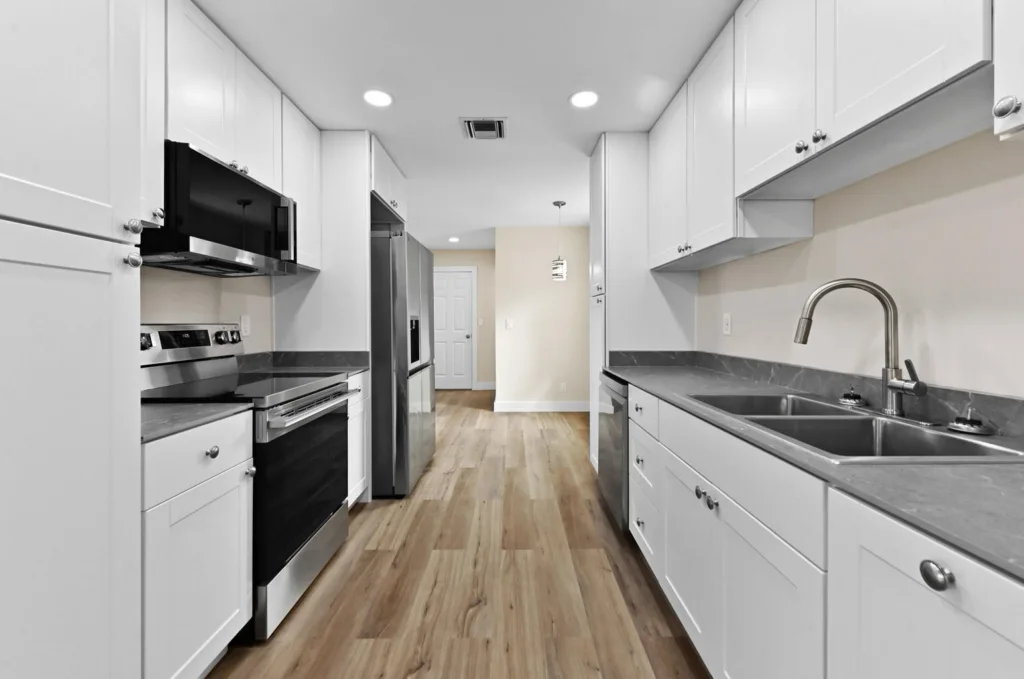 20261021192126515008000000-o 1 - Lake Worth Homes - Lake Worth Homes Bright galley kitchen with white cabinets, stainless steel appliances, and a double-basin sink along a gray countertop.