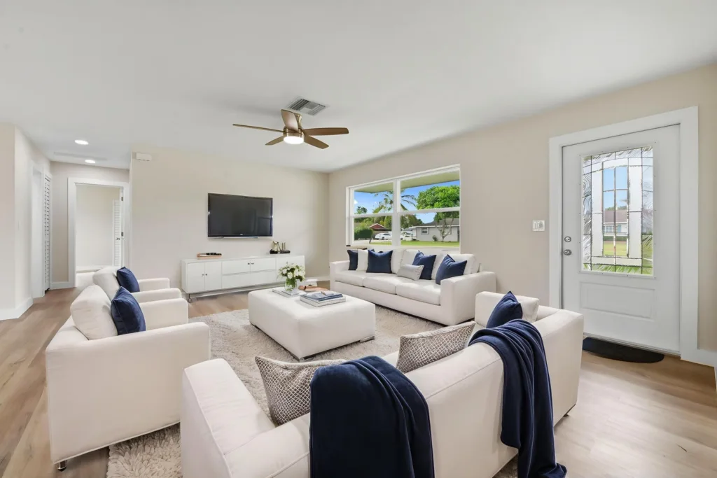 20261021191940478372000000-o 1 - Lake Worth Homes - Lake Worth Homes Bright modern living room with white sofas and navy throw blankets, a wall-mounted TV, and a large window overlooking greenery, creating an airy space.
