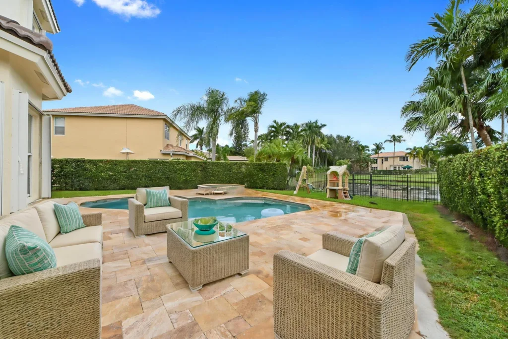 Backyard patio with wicker seating around a small pool, teal cushions, and palm trees under a bright blue sky.
