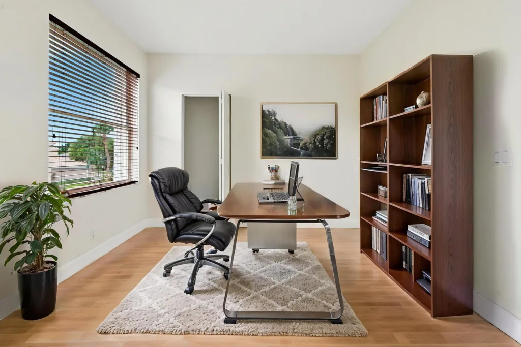 Modern home office with a wooden desk, black leather chair, and a tall bookshelf on the right; window with blinds on the left and a potted plant nearby.
