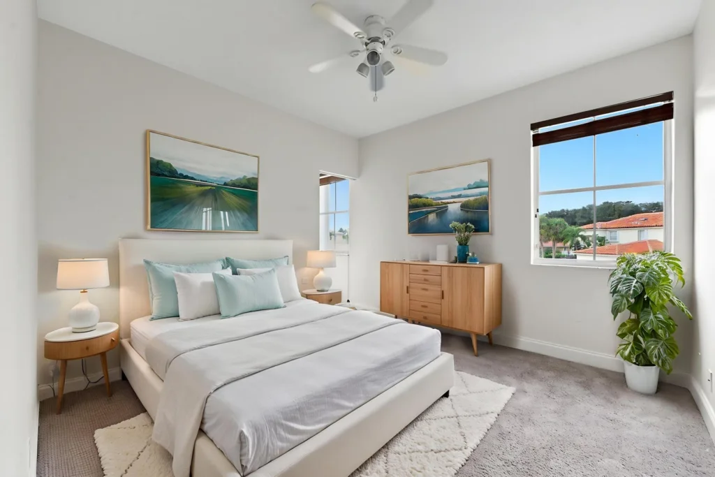 Bright modern bedroom with a white upholstered queen bed, light gray bedding, and teal accent pillows flanked by round wooden nightstands with lamps.