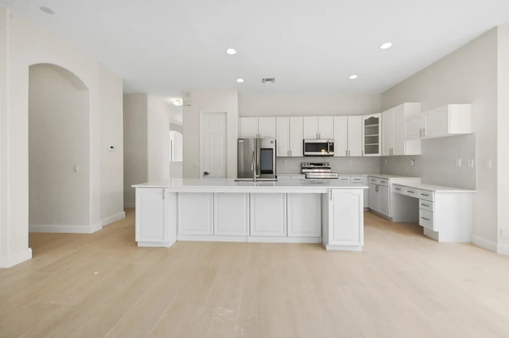 Bright, modern white kitchen with a large center island, stainless steel appliances, and white cabinetry on light wood floors.