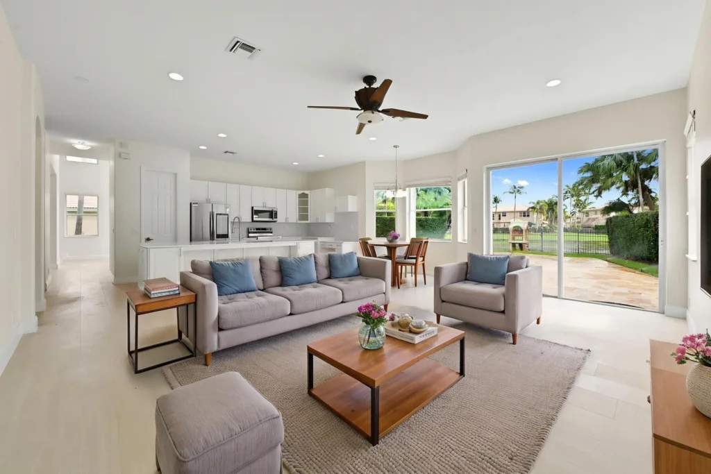 Open-concept living room with beige sofas, wooden coffee table, and large sliding glass doors opening to a sunny outdoor patio garden. Shows a connected kitchen and dining area in the background.