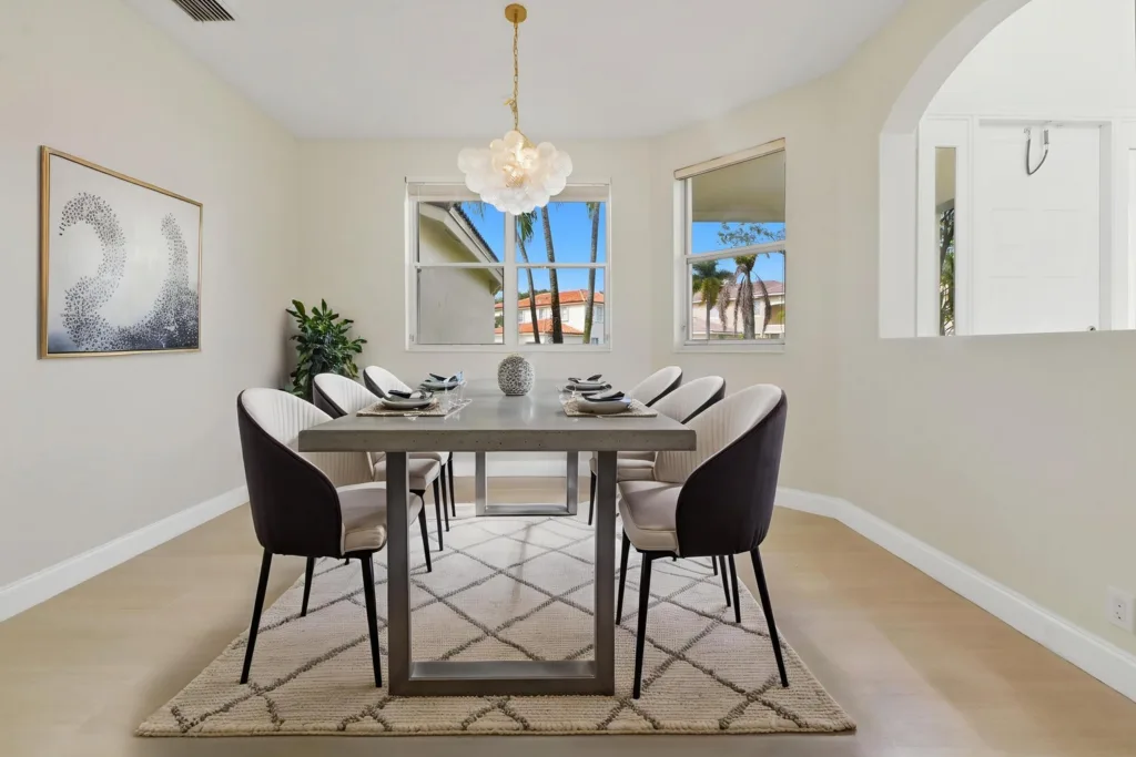 Modern dining room with a rectangular gray table, cushioned chairs, a decorative centerpiece, and a cloud-like pendant light; palm trees visible through the windows outside.