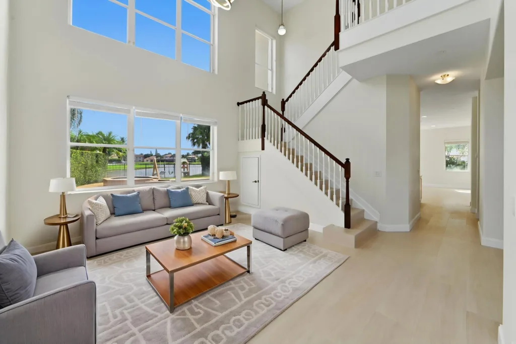 Bright two-story living room with large windows, gray sofa and armchairs, and a wooden coffee table beside a white-railed staircase.