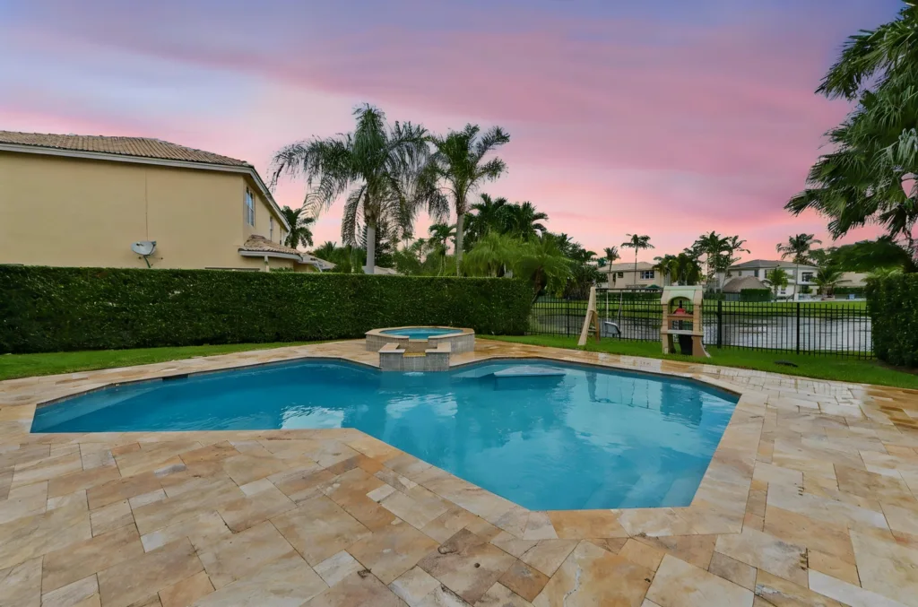 Backyard pool with an attached spa, tan stone patio, tall hedges, palm trees, and a pink sunset sky.