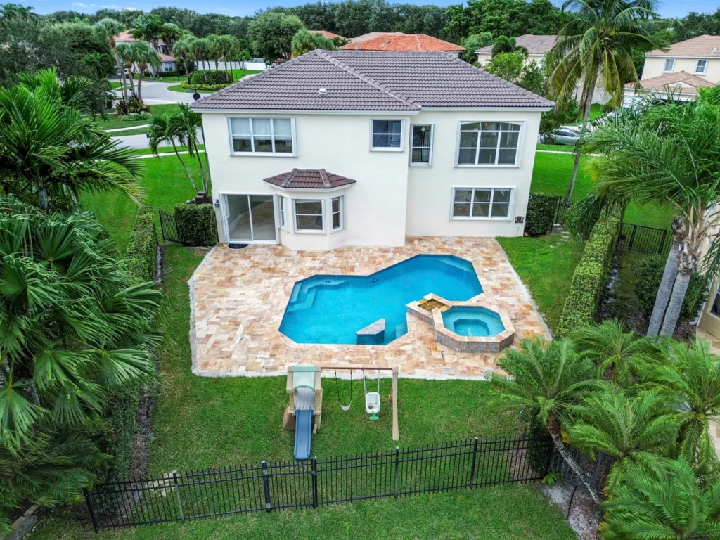 A two-story white house with a brown tiled roof, a stone-patio backyard, and a blue irregular-shaped swimming pool with an attached spa, surrounded by palm trees and a black fence.